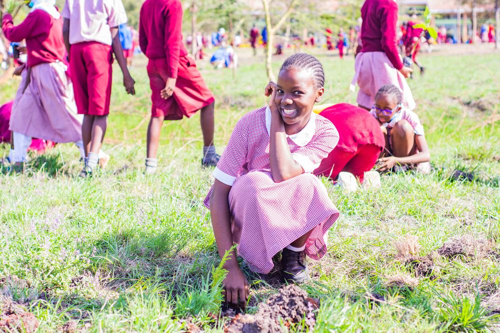 A smiling student kneeling beside a freshly planted seedling in a field